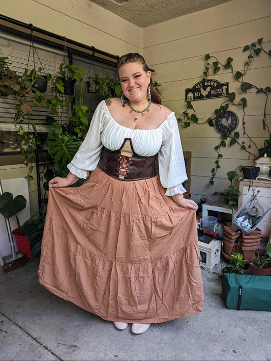 Alissa stands on a porch with green plants hanging from the window behind them. They have brown hair tied back are wearing a brown floor length skirt, a white top, and dark brown belt. They are holding their skirt out to the sides. 